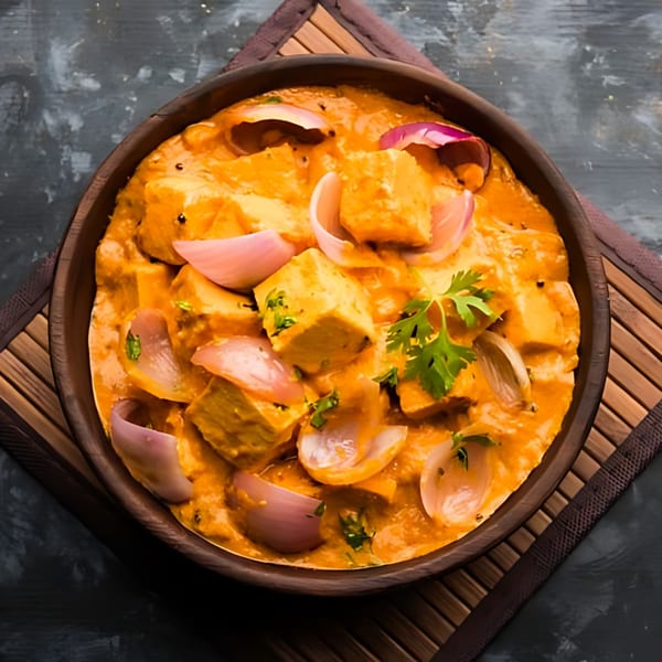 Paneer tikka masala with red onions and cilantro in a wooden bowl on a bamboo mat.