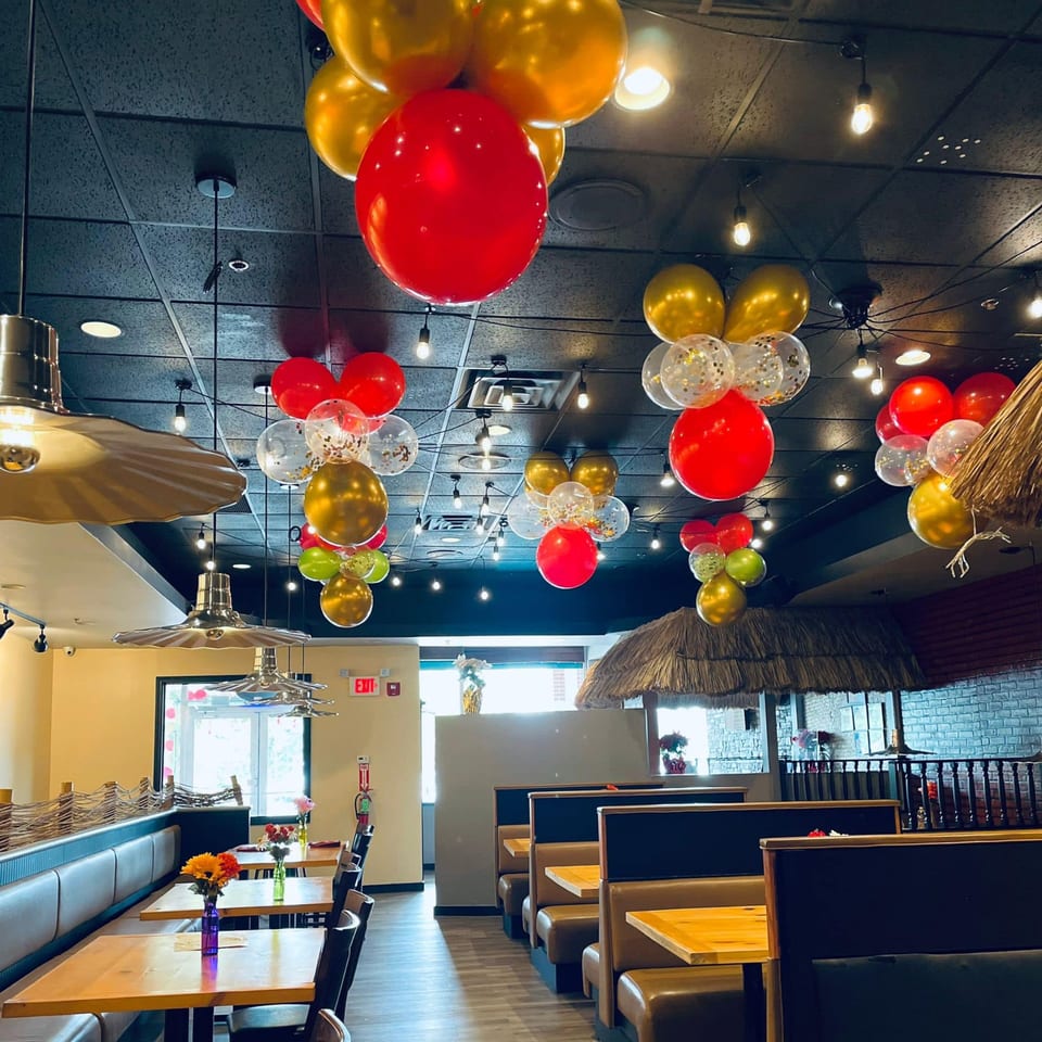 Restaurant interior with red and gold balloons hanging from the ceiling, booths, tables, and straw-covered structures.